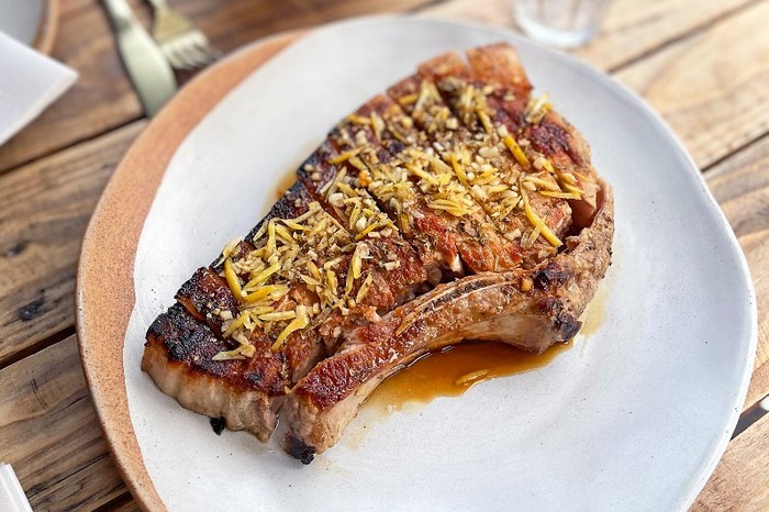 A large pork chop on a white plate served on a wooden table with white linen napkin