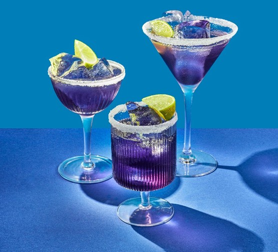 Three different shaped cocktail glasses filled with a purple drink, ice cubes and lime wedges on a blue background