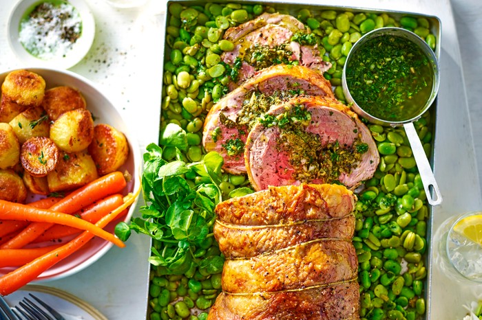 Rolled lamb joint in a roasting tin surrounded by green broad beans and a small pot of salsa verde, next to a bowl of carrots and potatoes