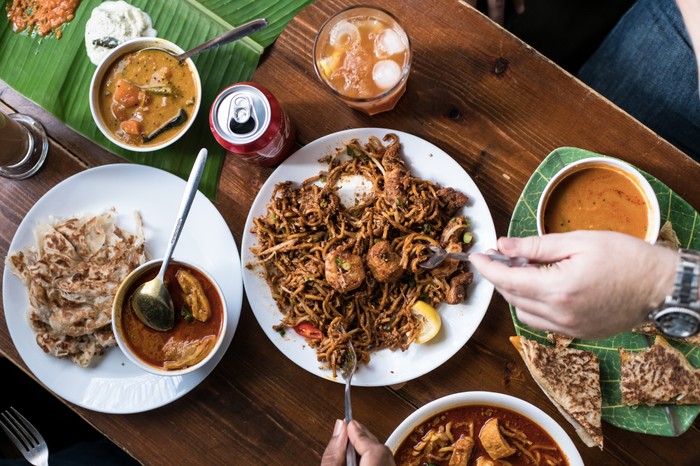 A selection of curries, roti and noodle dishes with hands tucking in to serve at Roti King