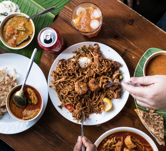 A selection of curries, roti and noodle dishes with hands tucking in to serve at Roti King