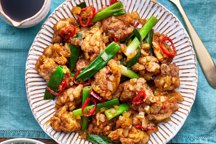 A plate of chicken with spring onions on a turquoise tablecloth with a bowl of rice and a spoon on the side