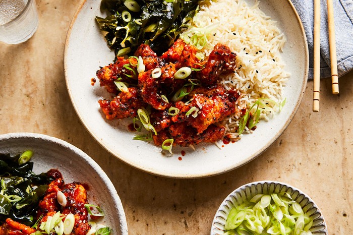 A plate of sesame chicken with rice, with a bowl of spring onions on the side and chopsticks