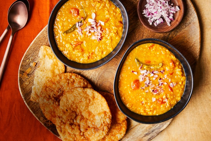 Two bowls of dhal next to a pile of pakwan bread and a ramekin of chopped onions