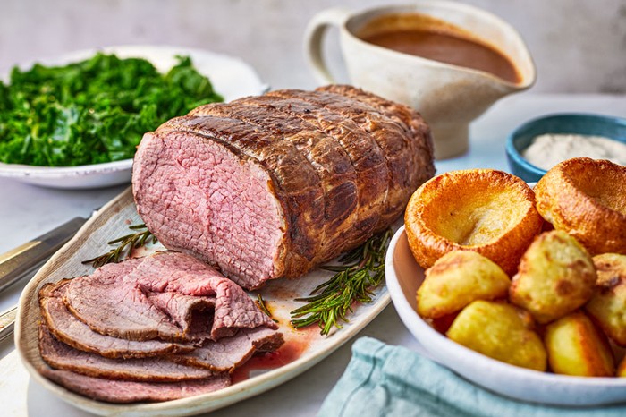 Sliced roast beef next to roast potatoes, yorkshire puddings and greens