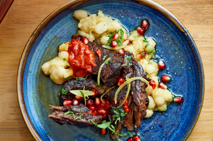 A blue plate with lamb neck on top of a bed of white beans, topped with pomegranate seeds and a red sauce.