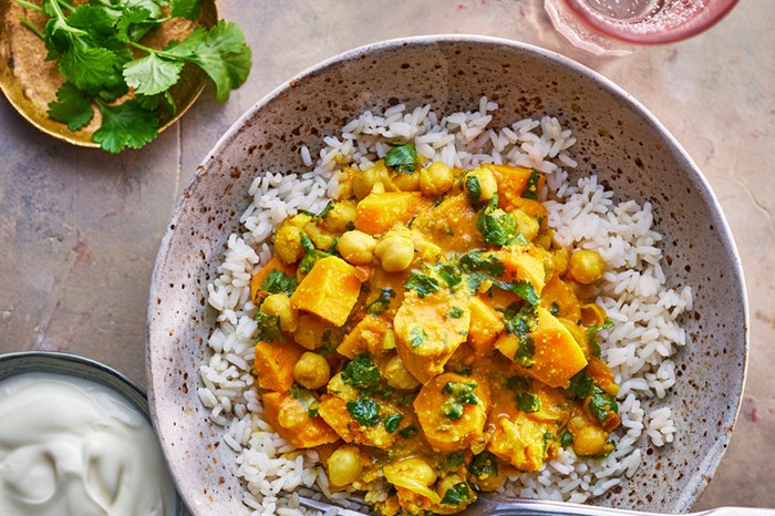 Bowl of sweet potato and chickpea curry on a bed of rice sprinkled with coriander, next to a bowl of yoghurt