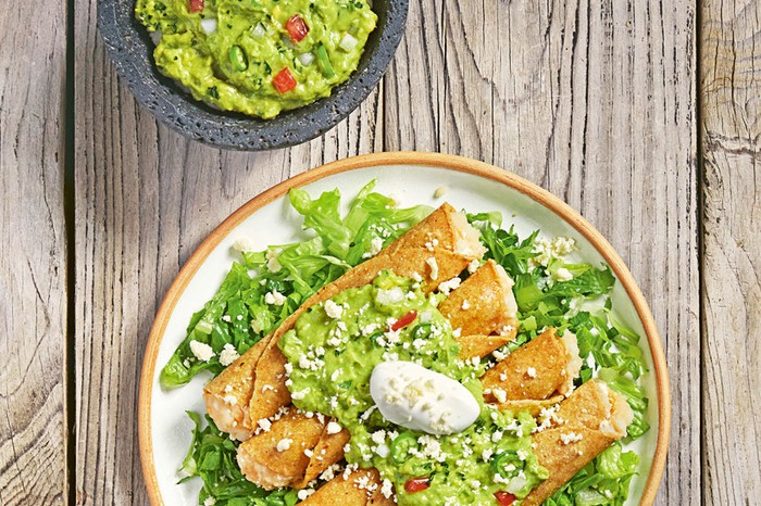 Rolled potato tacos on a bed of salad topped with guacamole and sour cream, next to a bowl of guacamole