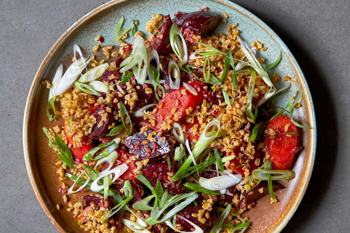 A plate of beetroot and golden freekeh topped with sliced spring onion on a grey background