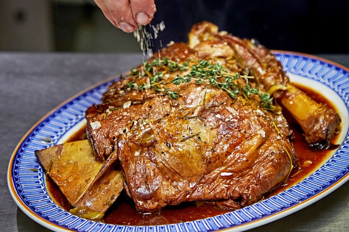 A whole braised shoulder of lamb on a serving plate with someone sprinkling salt on top