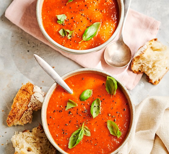 two bowls of red soup scattered with basil leaves and sat on folded linen napkins with chinks of crusty bread