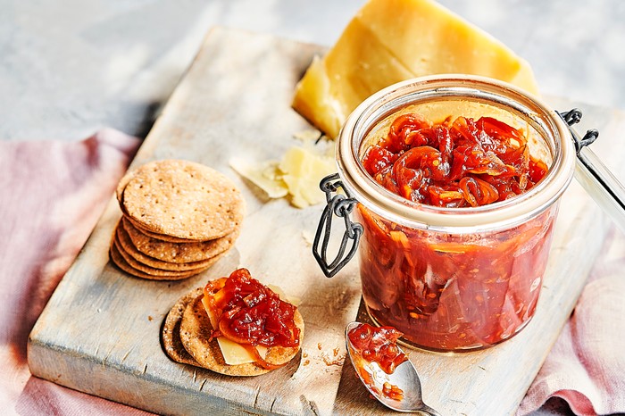 a jar of red tomato chutney on a wooden board with round crackers and a block of cheese