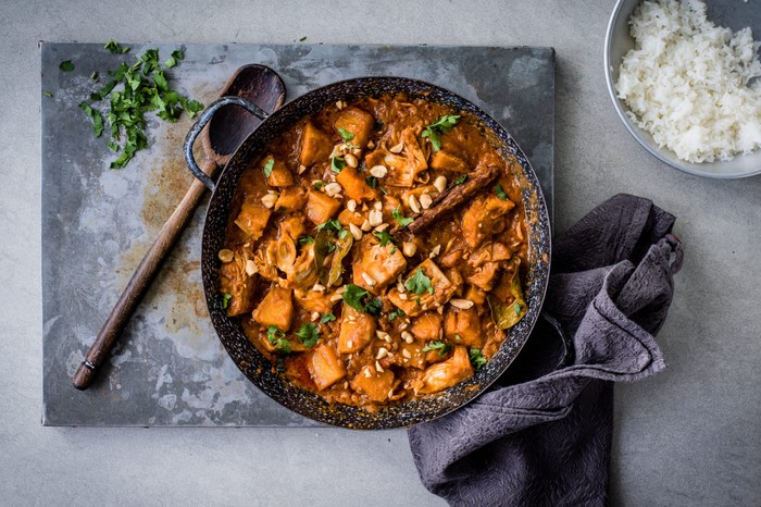 Skillet of vegan jackfruit curry next to a wooden spoon and tea towel