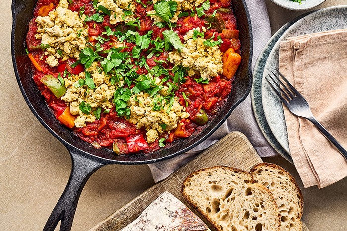 Vegan shakshuka topped with fresh parsley next to a board of bread and bowl of parsley and two plates with a tea towel