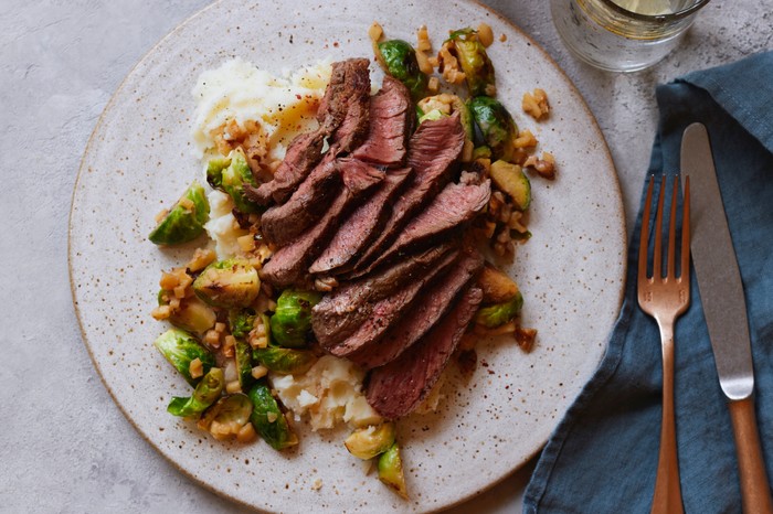 A speckled plate of thinly sliced seared meat with brussles sprouts, potatoes and walnuts