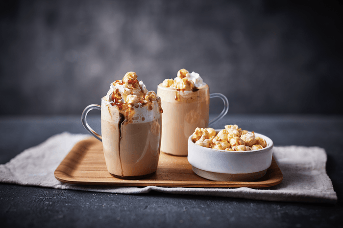 Two mugs of coffee topped with whipped cream and popcorn on a wooden tray alongside a bowl of popcorn