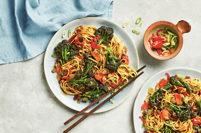 A white plate of broccoli, noodles and chilli with a pair of wooden chopsicks on a white background with a blue linen napkin