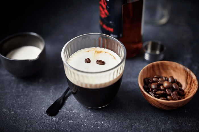 A ribbed glass of coffee with white cream on the top, garnished with two coffee beans next to a wooden bowl of coffee beans