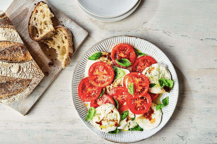 White plate filled with sliced tomatoes, sliced mozzarella and basil leaves, next to a board of sliced bread and some spare plates