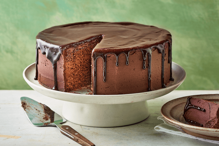 Chocolate cake with a glossy chocolate ganache dripping down the sides, sat on a white cake stand on a green background