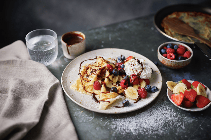 A plate of crepes on a stone surface topped with sliced fruit and a dusting of icing sugar