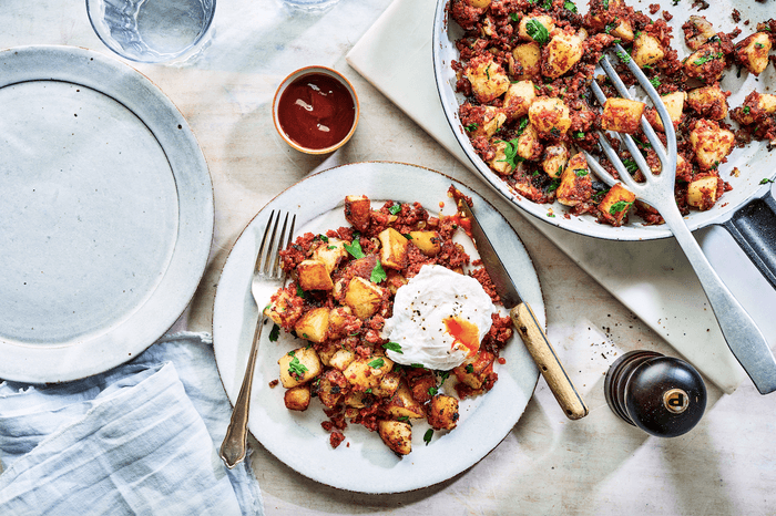 Corned beef hash Two white plates of golden fried potatoes topped with fried egg on a white wooden surface with a knife and fork and pot of brown sauce