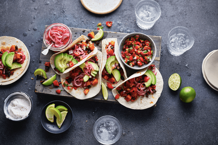 Four tacos on a wooden board with small bowls of salsa and pink pickled onions with wedges of lime on a grey stone background