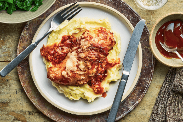 A plate of mash topped with hunter's chicken on a wooden background, with a small pot of BBQ sauce on the side