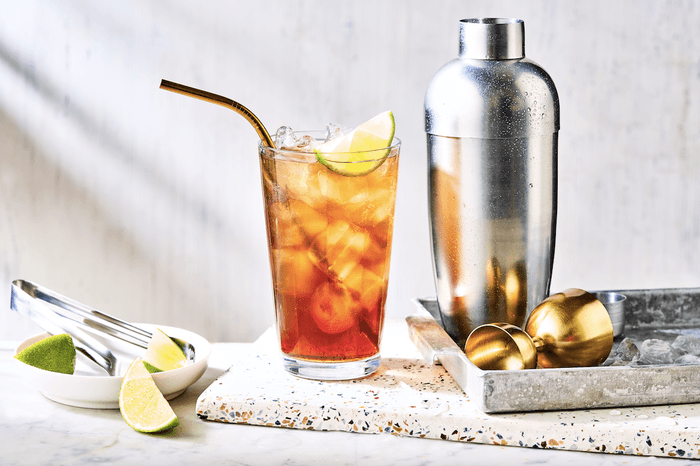 an amber-cooured cocktail in a tall glass next to a silver cocktail shaker with wedges of lime