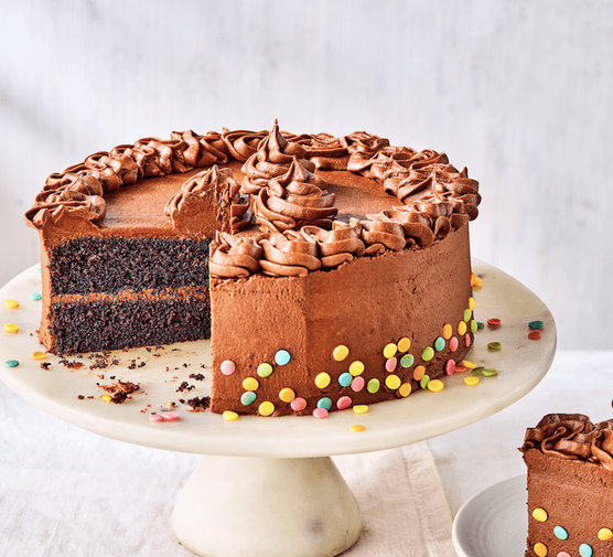 A chocolate frosted cake on a white cake stand with a slice cut out of it in front of a white background